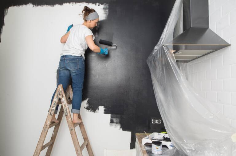 Donna su una scala che tinteggia con il rullo la parete della cucina con pittura lavabile scura