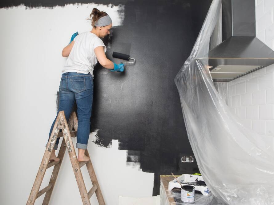 Donna su una scala che tinteggia con il rullo la parete della cucina con pittura lavabile scura